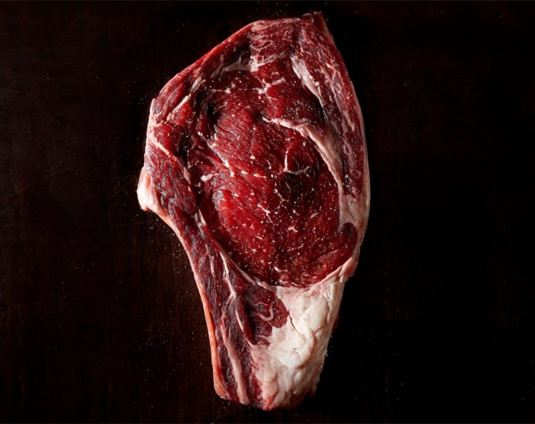 Close-up of a raw bone-in ribeye steak on a dark background, photographed by OMS Photo, expert commercial food and protein photographer.