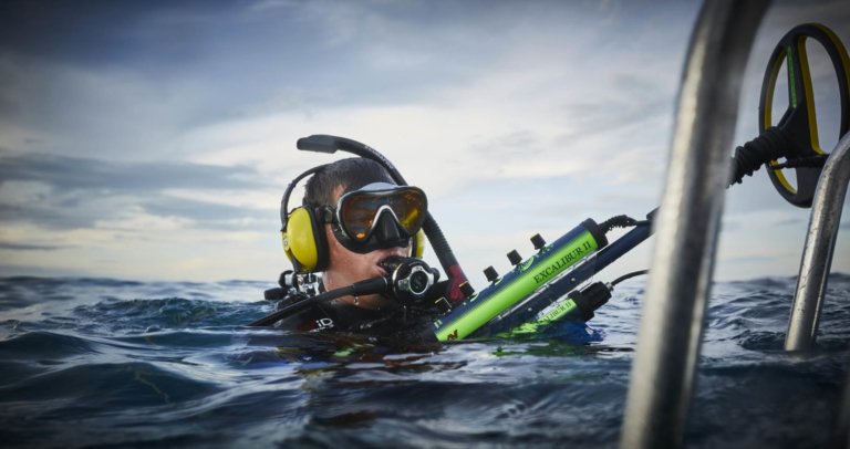 A diver ready to dive holding a metal detector at the surface