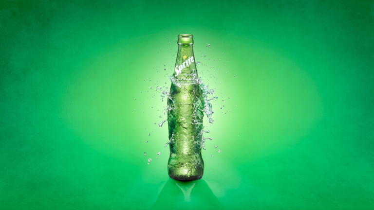 Glass beverage Sprite bottle with dramatic water splash on green background captured using high-speed liquid photography by commercial drink photographer OMS Photo