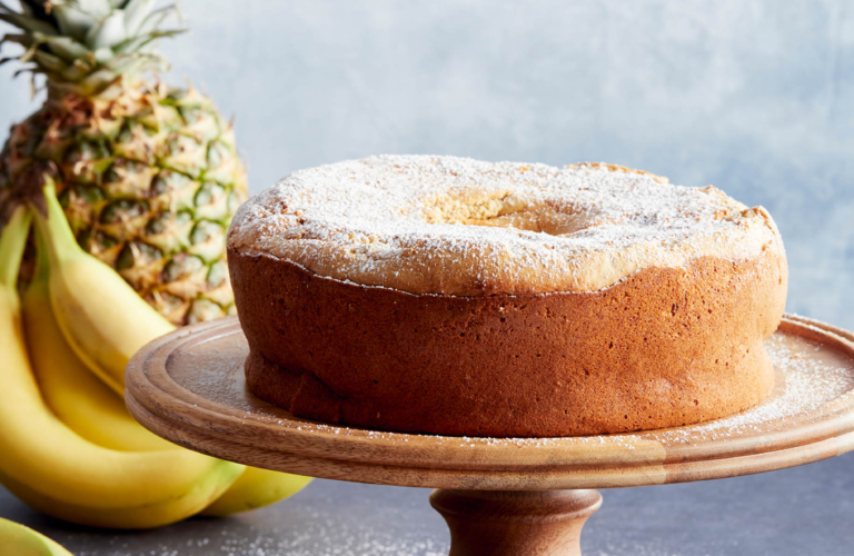 Whole pineapple-banana bundt cake dusted with powdered sugar on a wooden cake stand, with fresh pineapple and bananas styled in the background.