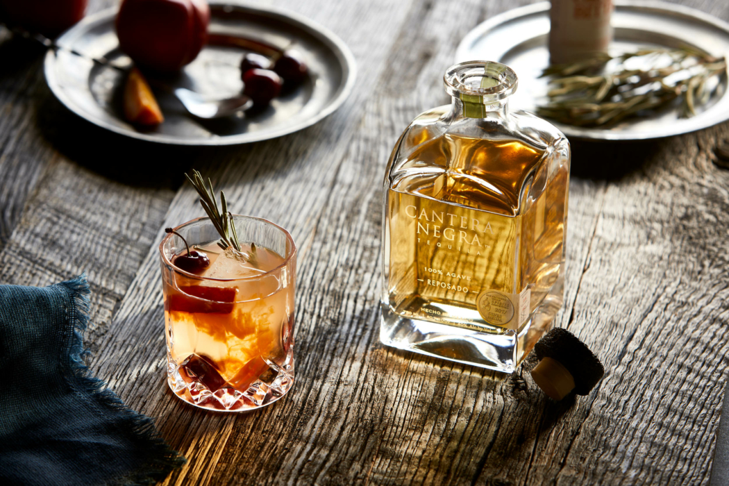 Drink photography - a bottle of cantera negra with an orange-tinted cocktail with a cherry and rosemary garnish