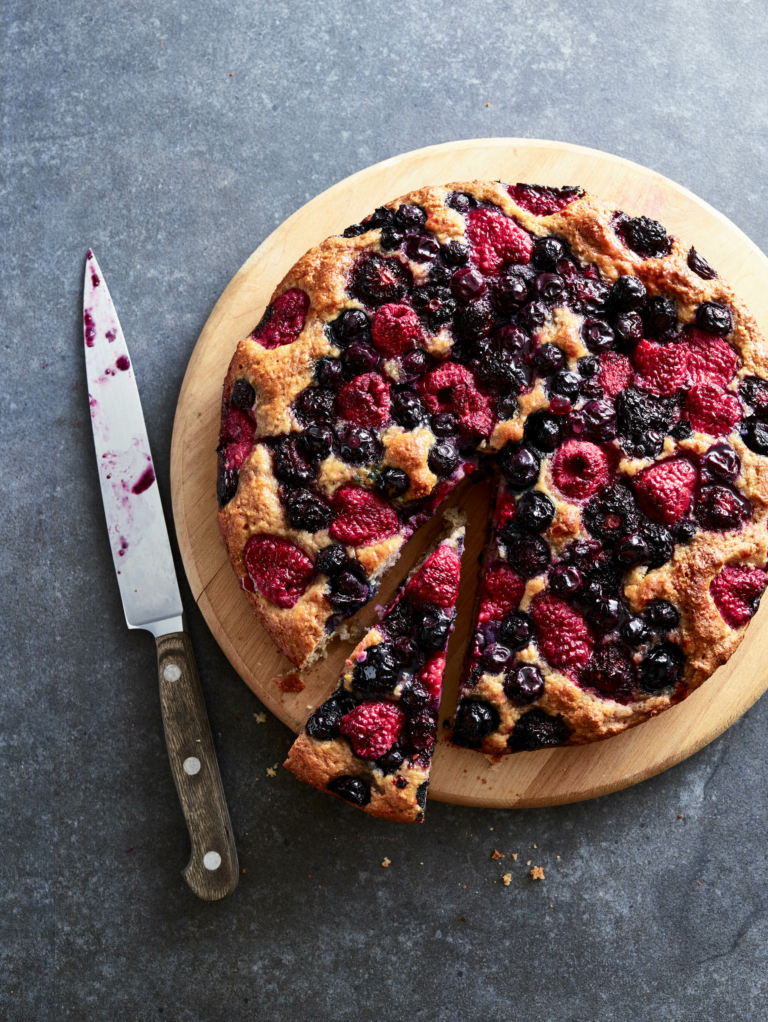 Overhead shot of a rustic mixed berry cake on a wooden board with a slice removed and a berry-stained knife beside it.