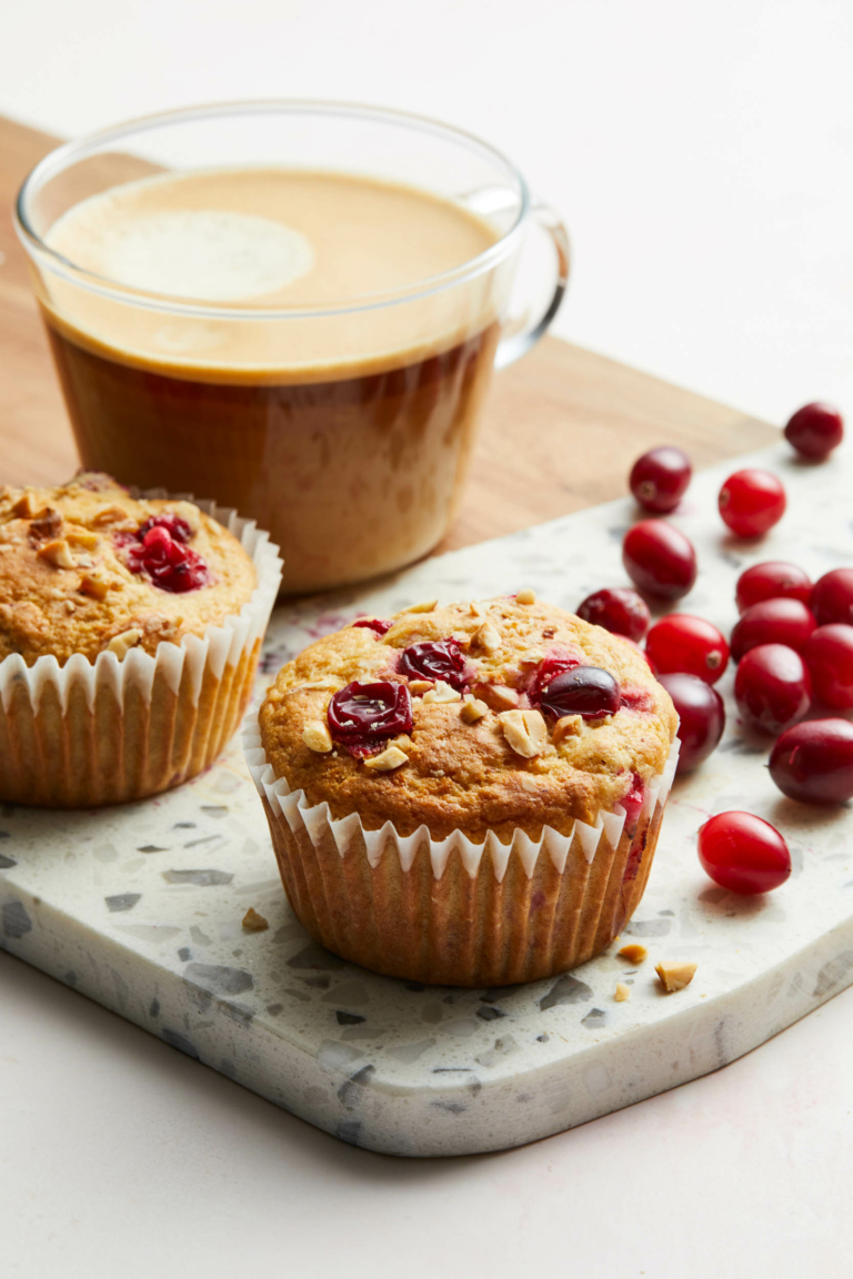 Cranberry muffins on terrazzo board beside a glass mug of latte and fresh cranberries, photographed in soft morning light.