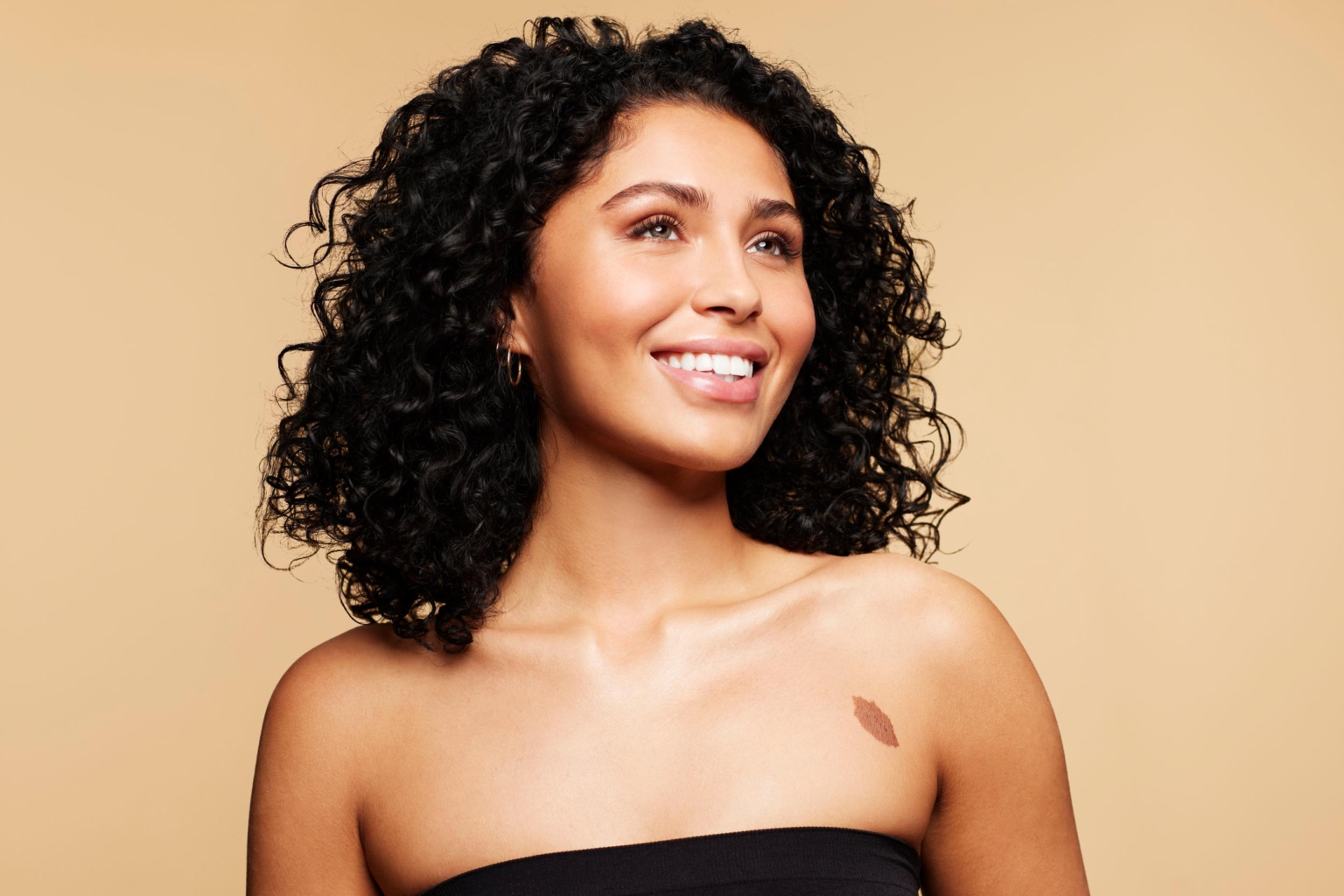 Woman with natural curly hair smiling and looking off-camera against a neutral beige background, beauty photography by Monty Milburn
