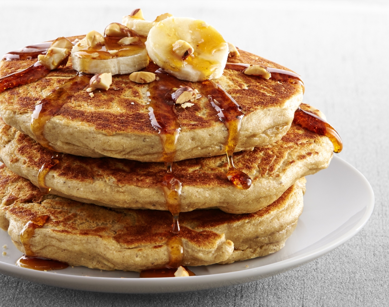 Stack of pancakes topped with banana slices, chopped walnuts, and syrup drizzling down the sides on a white plate.