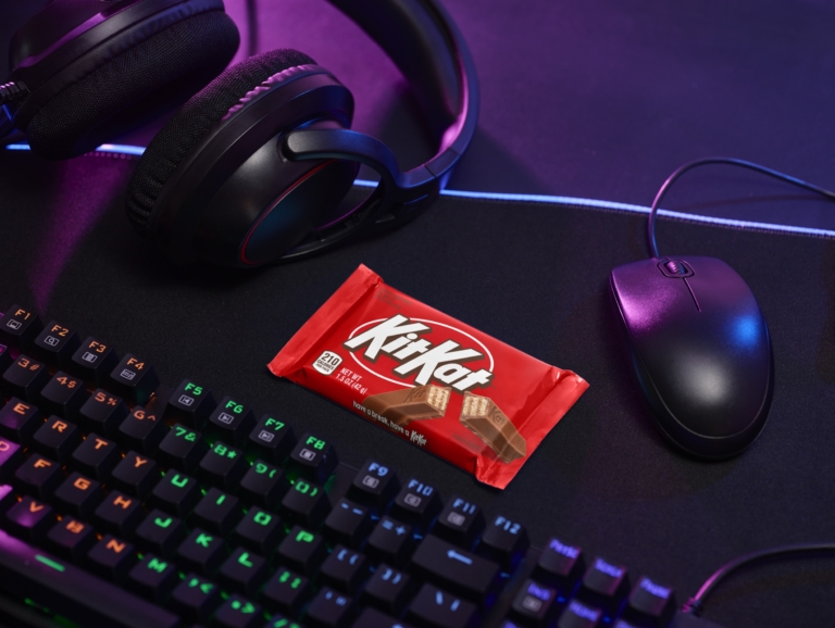KitKat chocolate bar placed on a gaming desk with RGB keyboard, headphones, and mouse under purple lighting.