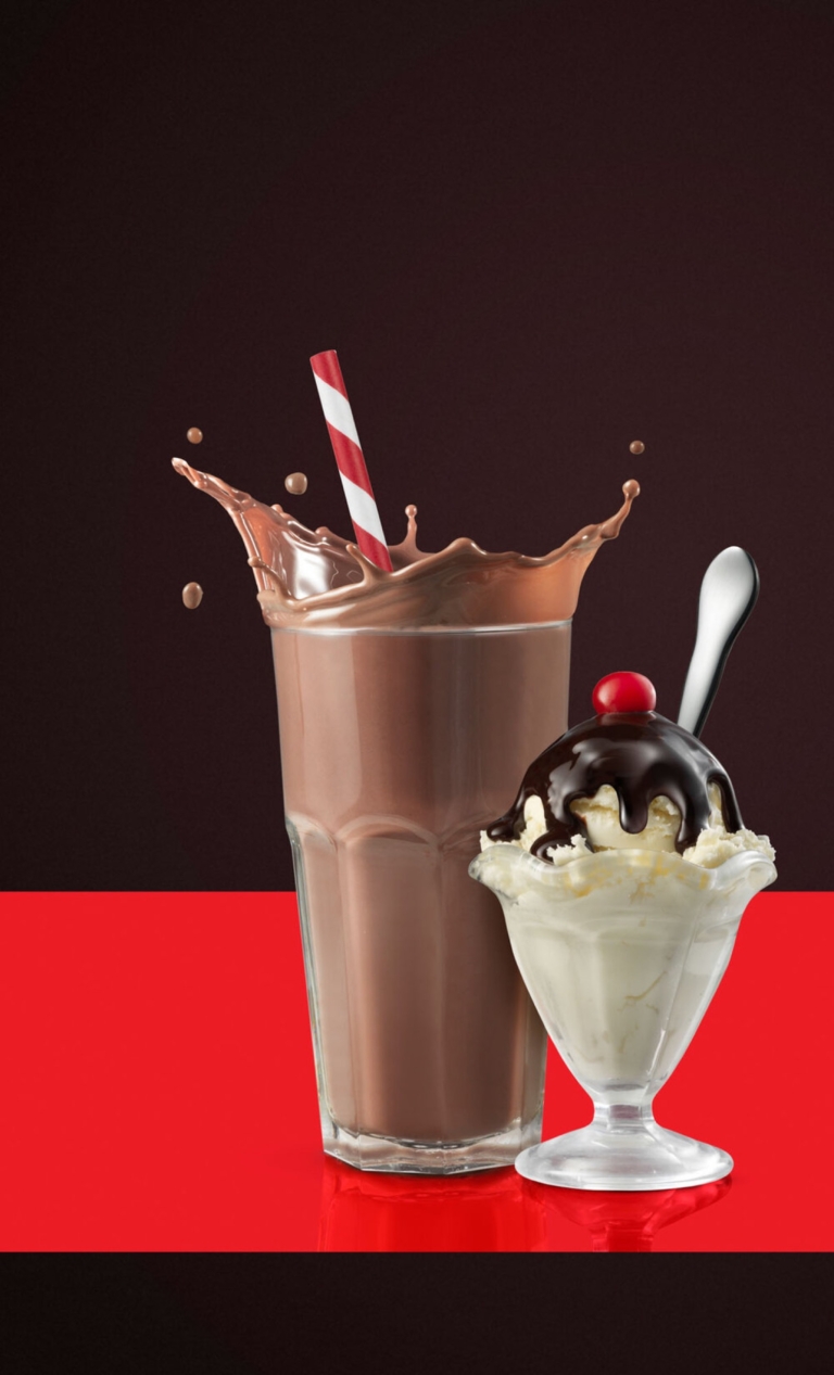 Tall chocolate milkshake splashing upward beside a vanilla sundae topped with chocolate sauce and a cherry.