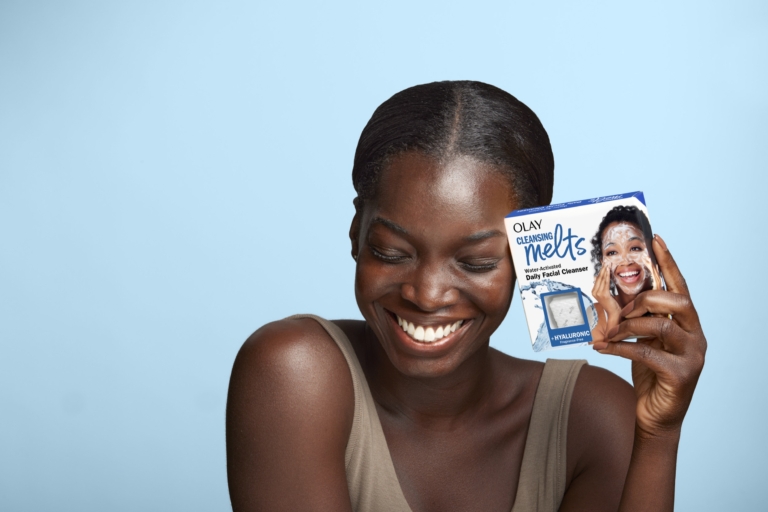 Smiling Black woman holding an Olay skincare product against a light blue background