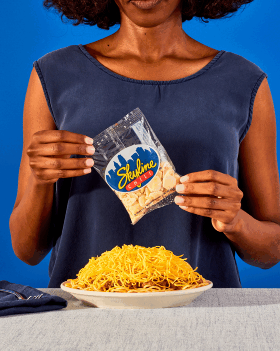 Woman holding a bag of Skyline Chili oyster crackers over a plate of three way spaghetti topped with shredded cheese against a blue background