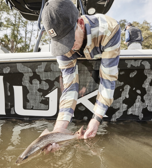 Man wearing HUK gear catching a fish over the side of a boat, captured in action for outdoor adventure and fishing lifestyle photography by OMS Photography