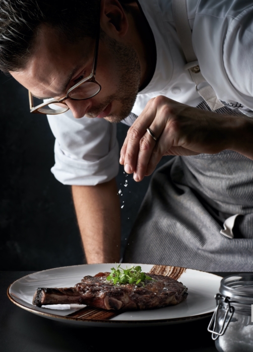 a man in glasses and a chef's uniform sprinkling salt on a plate of food, chef and restaurant photography by OMS Photographer