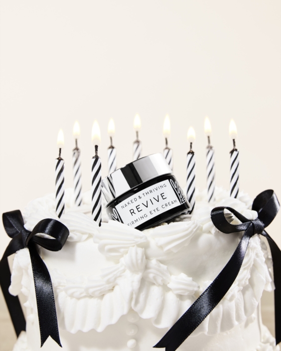 A Revive skincare jar sits on top of a white birthday cake surrounded by lit striped candles.
