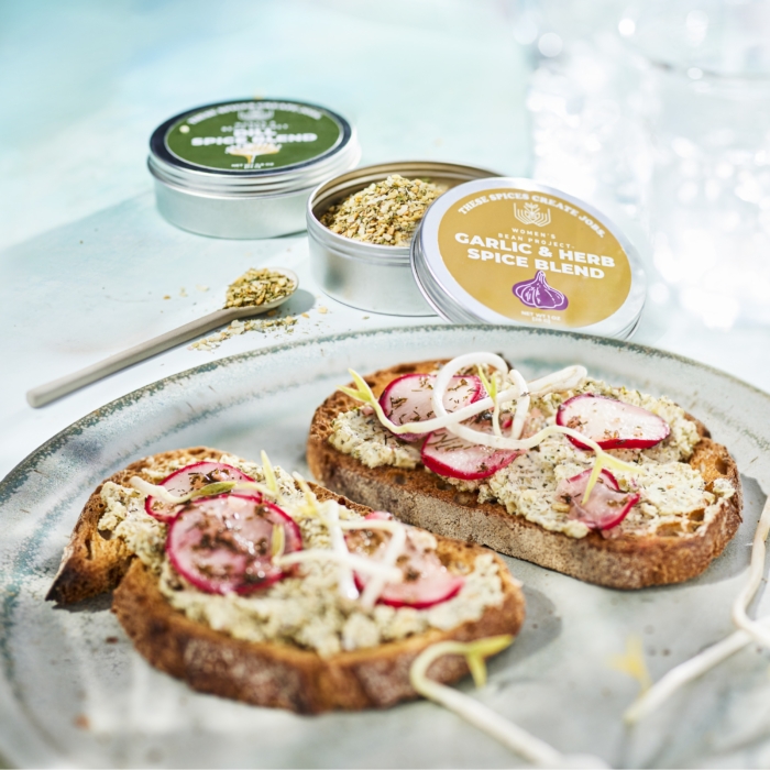Two slices of radish toast topped with herbed spread and sliced onions, photographed with open tins of Women’s Bean Project Garlic & Onion Spice Blend and Dill Spice Blend on a bright tabletop.