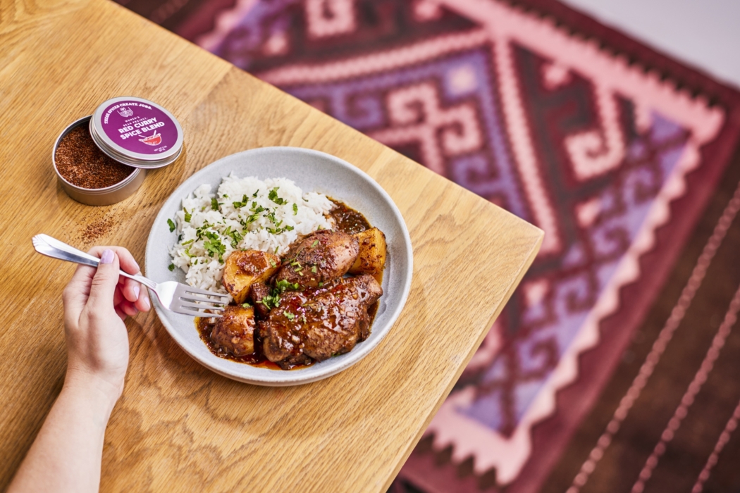 Bowl of glazed chicken with rice on a wooden table, photographed alongside an open tin of Women’s Bean Project Purple Pepper Spice Blend, with a patterned rug in the background.