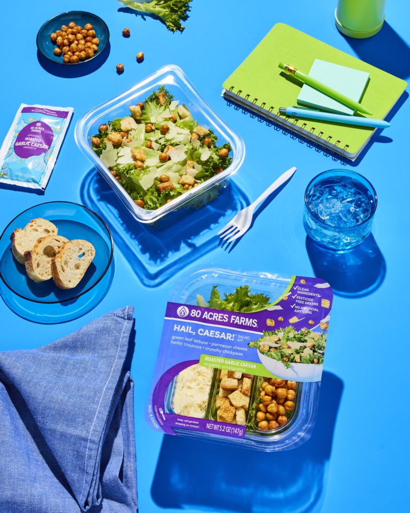 Overhead image of a Caesar salad kit, plated salad, drink, bread, and office supplies arranged on a bright blue background.