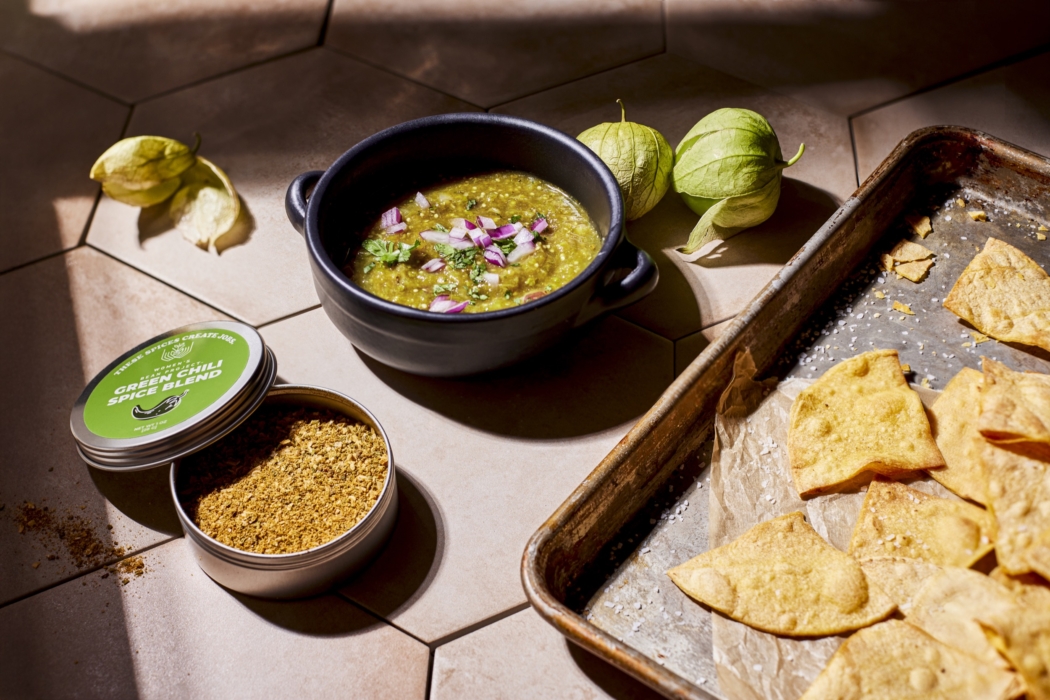 Bowl of salsa verde garnished with onions and cilantro next to tomatillos, baked tortilla chips on a tray, and an open tin of Women’s Bean Project Jalapeño Lime Spice Blend.