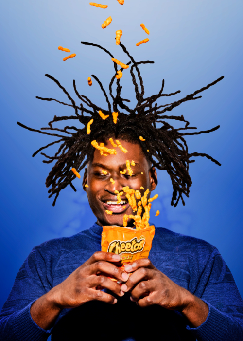 Vibrant portrait of a man joyfully opening a Cheetos bag as cheese puffs explode upward into his hair against a blue gradient background.