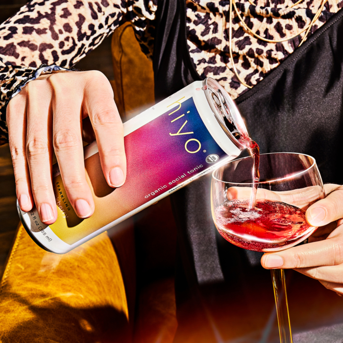 Close-up of a hand pouring a can of Hiyo organic social tonic into a glass, with vibrant lighting and a stylish outfit.