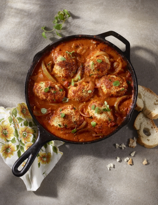 Overhead shot of chicken simmering in a tomato cream sauce inside a cast-iron skillet, served with fresh herbs and sliced bread on the side.
