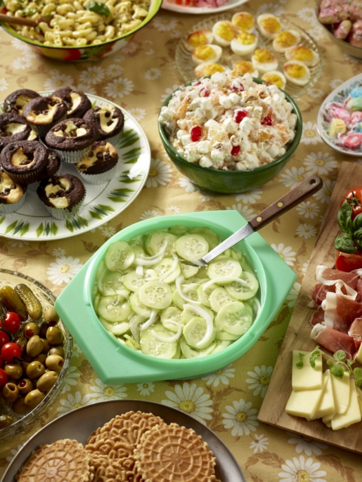 Overhead shot of a vintage-style potluck table with cucumber salad, deviled eggs, popcorn salad, olives, cheese skewers, and pastel candies.