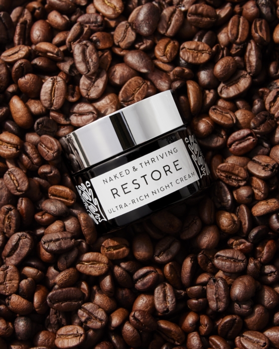 Naked & Thriving Restore moisturizer jar surrounded by coffee beans on dark background