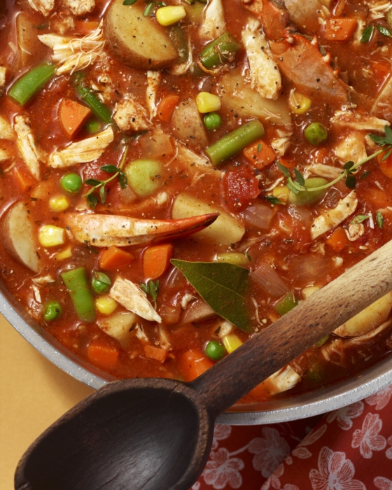 Close-up of a pot of chicken vegetable stew with carrots, potatoes, green beans, peas, corn, and herbs, with a wooden spoon resting beside it.