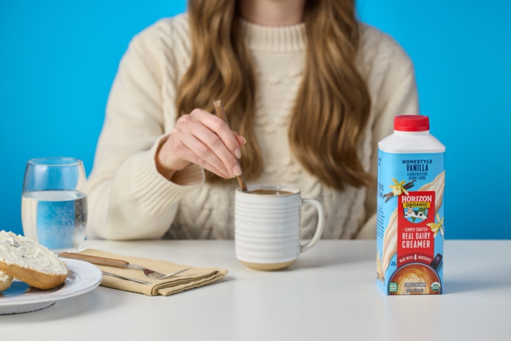 Hand pouring Horizon Organic coffee creamer into glass mug of coffee against blue background.