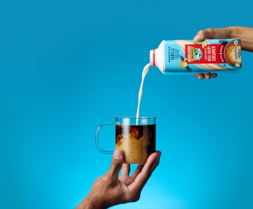Hand pouring Horizon Organic coffee creamer into glass mug of coffee against blue background.