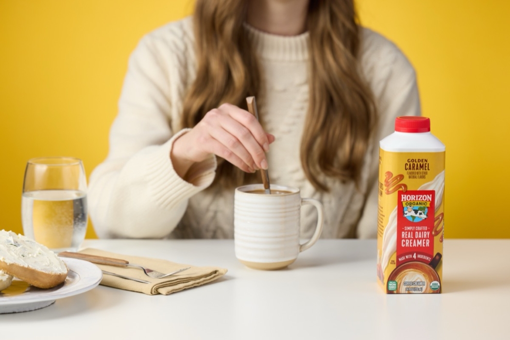 Hand pouring Horizon Organic coffee creamer into glass mug of coffee against yellow background.