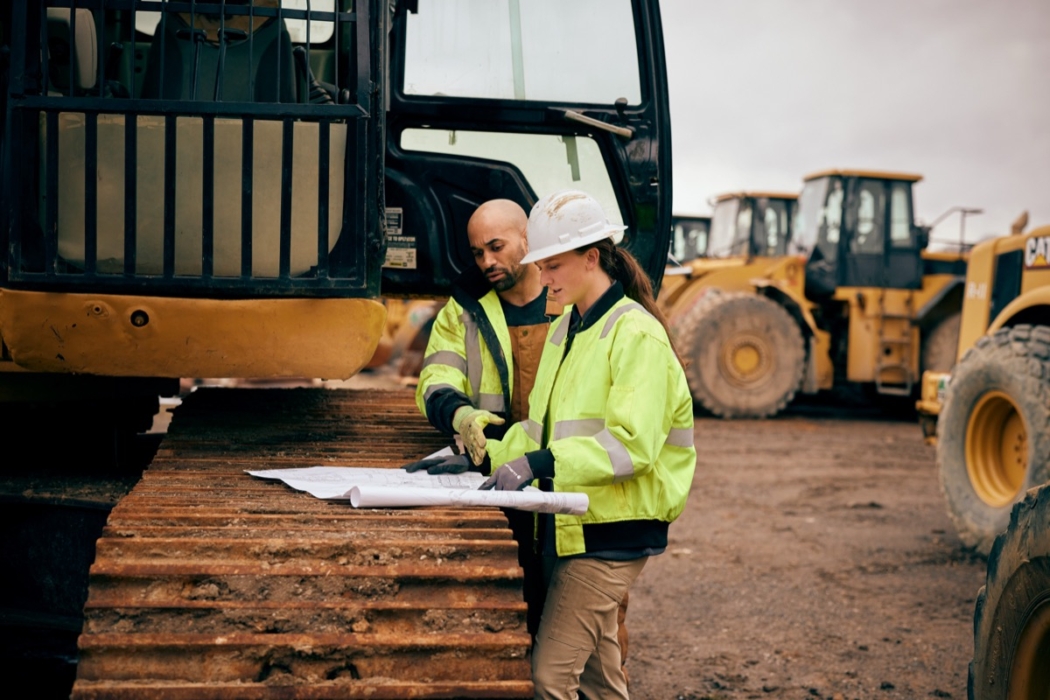 Two construction workers in hard hats reviewing blueprints on excavator tread beside heavy equipment lineup