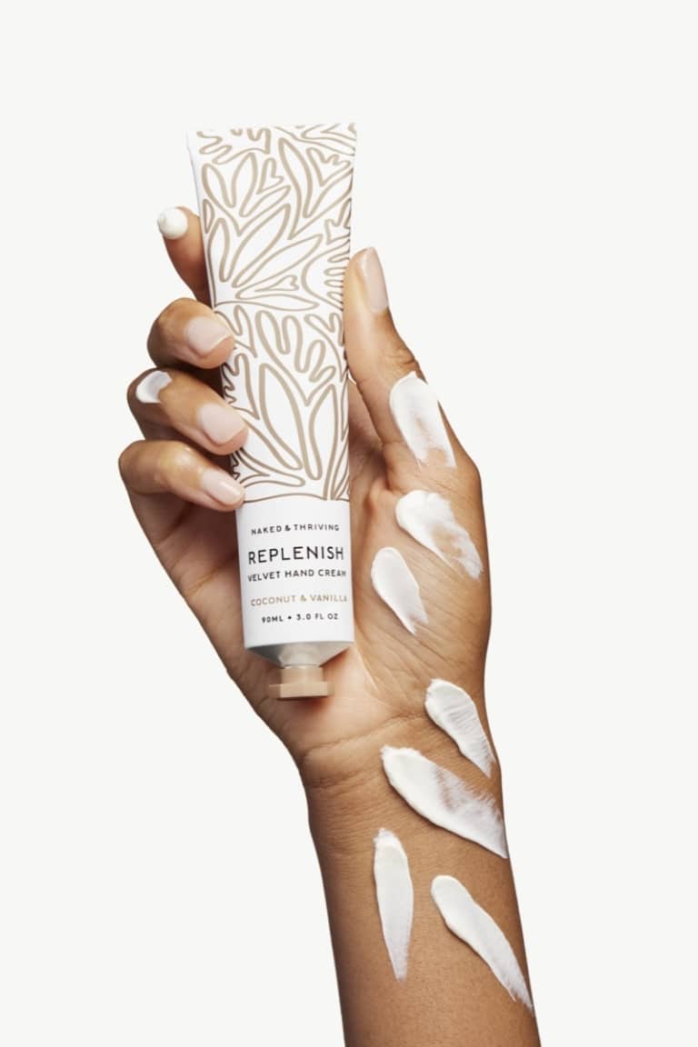 Hand holding Naked & Thriving Replenish hand cream tube with cream swirls on warm neutral background