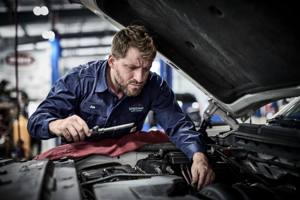 Male auto mechanic in blue uniform leaning over engine bay inspecting vehicle components in automotive shop