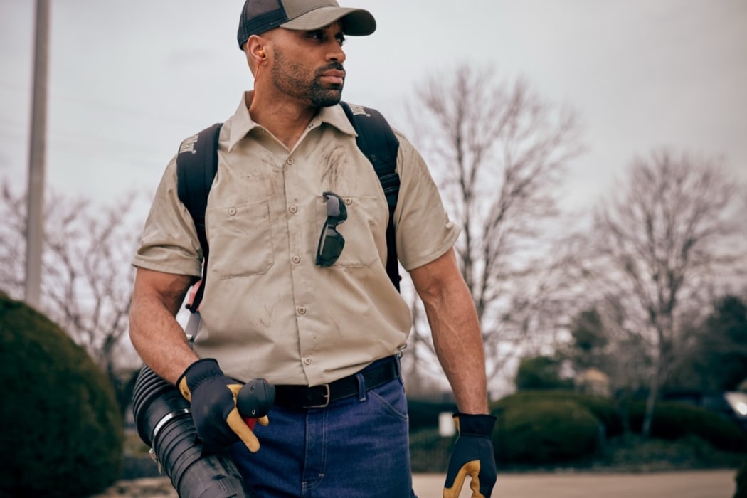 Male landscaper in tan work shirt and gloves carrying a backpack leaf blower at an outdoor job site