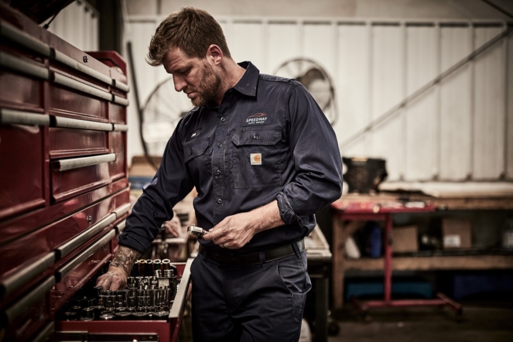 Male mechanic in dark navy Carhartt work shirt examining tools at a workbench in an industrial shop