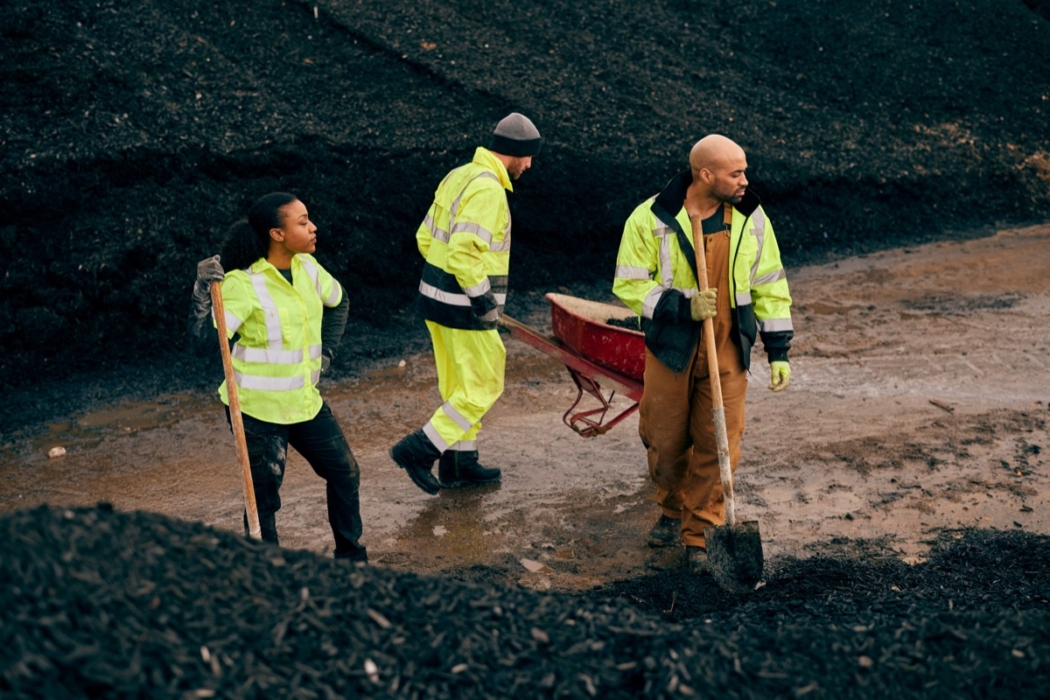 hree workers in high-visibility jackets shoveling and moving mulch with wheelbarrow at composting site