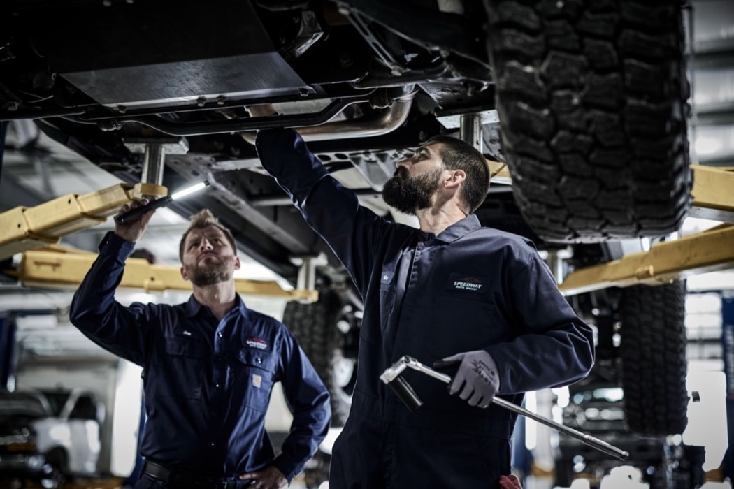 Two male mechanics in navy uniforms working underneath a lifted vehicle in an automotive shop