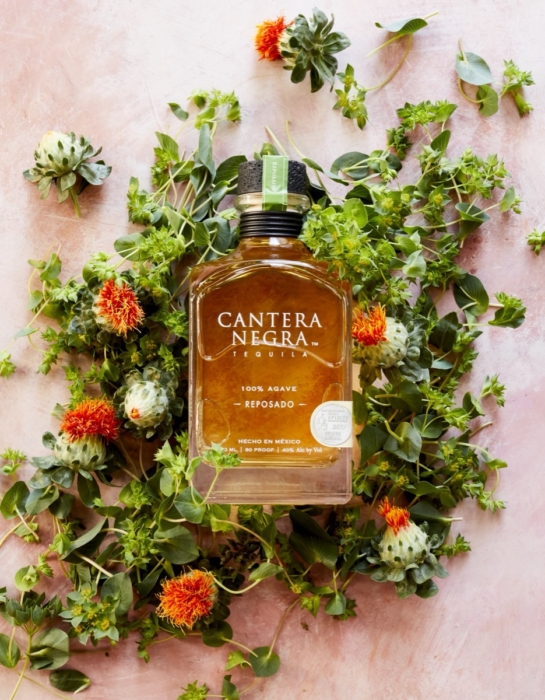 Cantera Negra Reposado Tequila bottle surrounded by safflower blooms and greenery in overhead flat lay photography