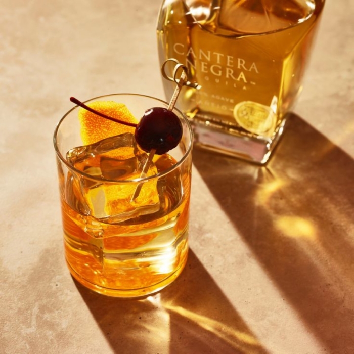 Amber tequila old fashioned cocktail with cherry and orange peel garnish beside Cantera Negra Extra Anejo bottle