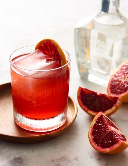Red blood orange tequila cocktail with citrus wheel garnish on wooden coaster beside fresh blood orange wedges and tequila bottle