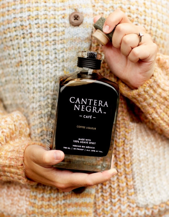 Close-up of hands opening Cantera Negra Cafe Coffee Liqueur bottle cork top while wearing cream knit sweater