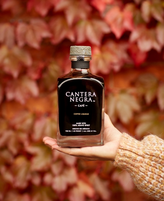 Hand holding Cantera Negra Cafe Coffee Liqueur bottle against blurred red autumn ivy leaves background