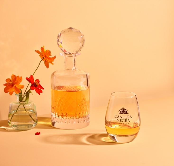 Cantera Negra Tequila poured into crystal decanter and branded stemless glass on warm peach background with fresh flowers
