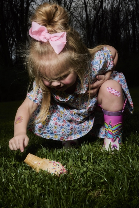 Young child in a dress picking up a dropped ice cream cone on grass outdoors