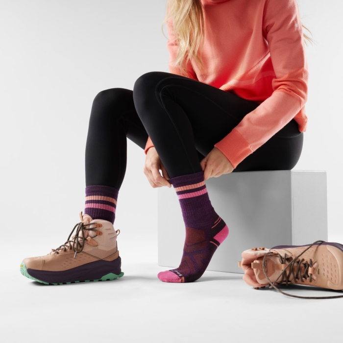 Woman seated pulling up pink striped performance sock with hiking boots nearby, on-model sock and footwear product photography by OMS Photo