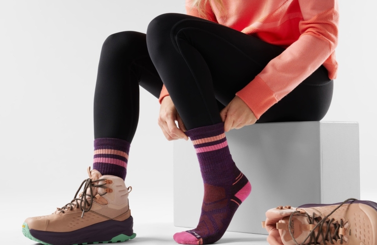 Woman seated pulling up pink striped performance sock with hiking boots nearby, on-model sock and footwear product photography by OMS Photo
