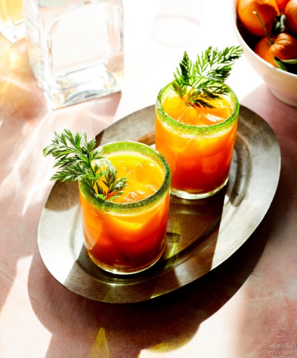 Two orange tequila cocktails with green sugar rims and fresh herb garnish served on metal tray with citrus in background
