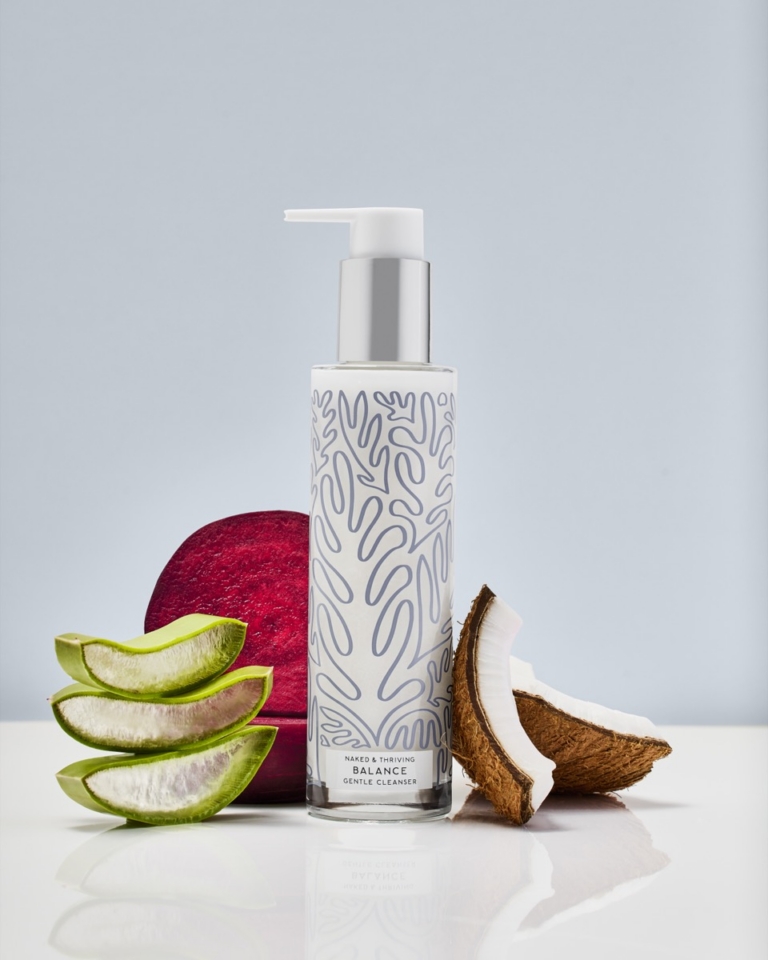 Naked & Thriving Balance Gentle Cleanser pump bottle surrounded by fresh sliced aloe vera leaves, a halved beetroot, and a cracked open coconut on a white reflective surface with soft grey background, communicating key natural skincare ingredients