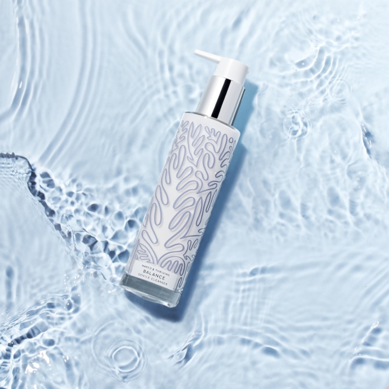 Overhead flatlay of Naked & Thriving Balance Gentle Cleanser pump bottle floating in shallow rippling water on a pale blue surface with concentric water ring reflections visible around the bottle