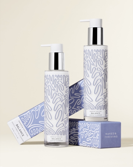 Two Naked & Thriving Balance Gentle Cleanser pump bottles of different sizes arranged with the periwinkle blue retail box and a branded Naked & Thriving product cube against a warm cream background, editorial product styling