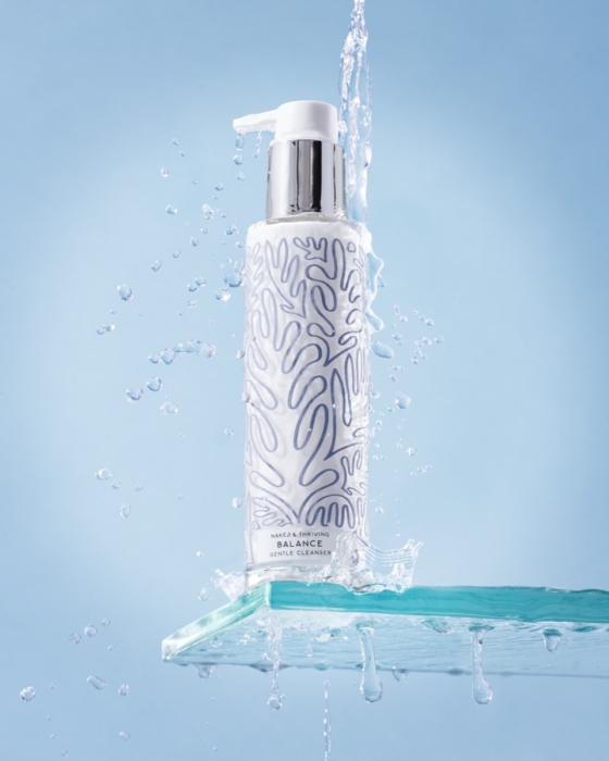 Naked & Thriving Balance Gentle Cleanser standing upright on a clear glass shelf with water splashing dramatically around and over it against a light blue background, water droplets suspended mid-air